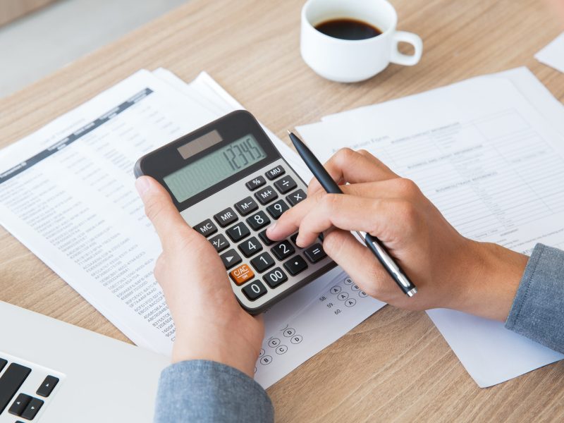 Unrecognizable businessman using calculator to find out income and outcome. He counting company finances. Male hands with pen pushing button at desk. Investment or paperwork concept